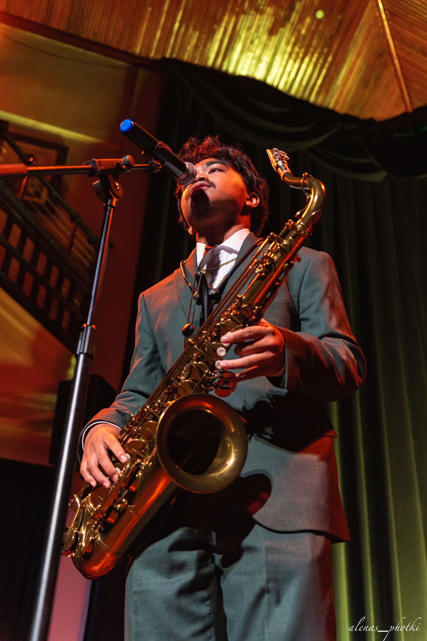 A saxophone player in a gray suit performs on stage under warm lighting at a concert venue.