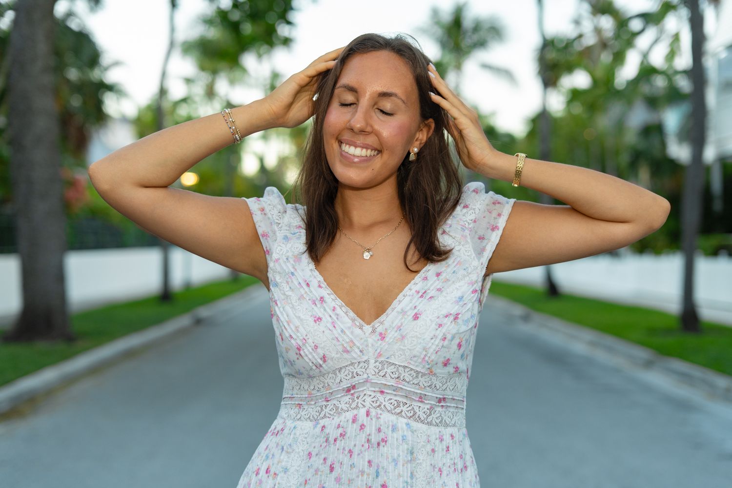 A woman in a white floral dress poses joyfully outdoors with palm trees in the background.