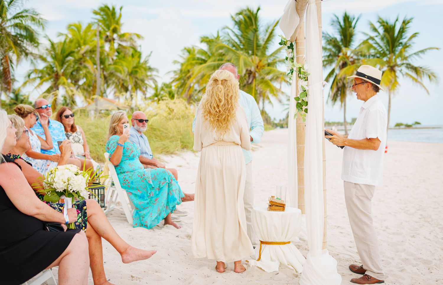 A beach wedding ceremony with guests in bright colors and palm trees swaying in the background.