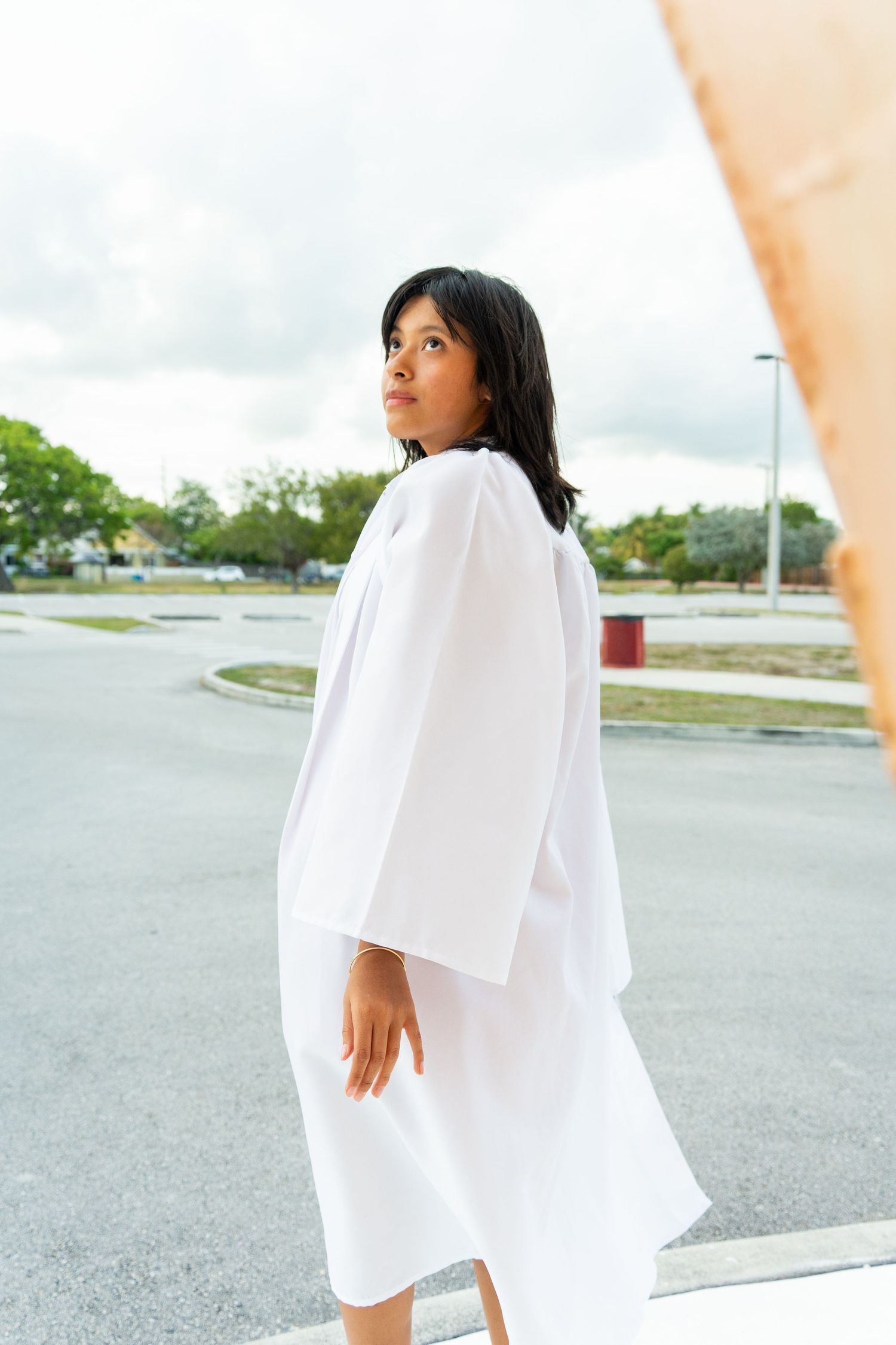 Young woman in elegant white oversized dress poses outdoors in parking lot with cloudy sky background.