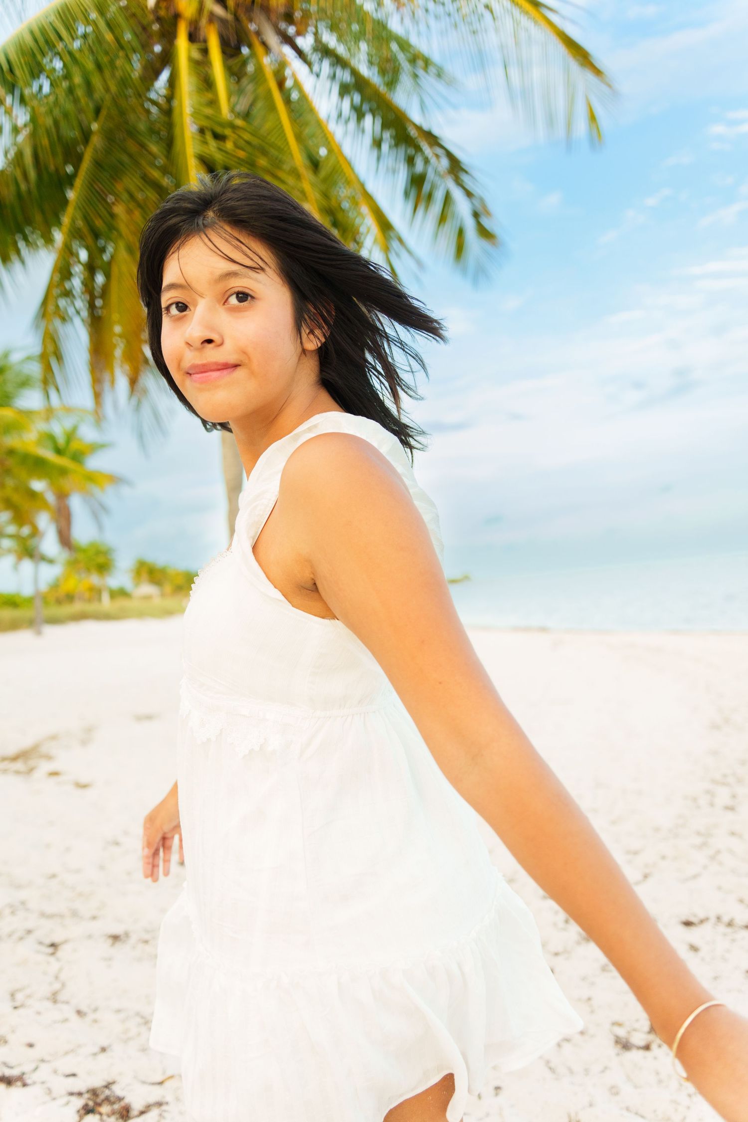 Young woman in white sundress smiling on tropical beach with palm trees and blue sky.