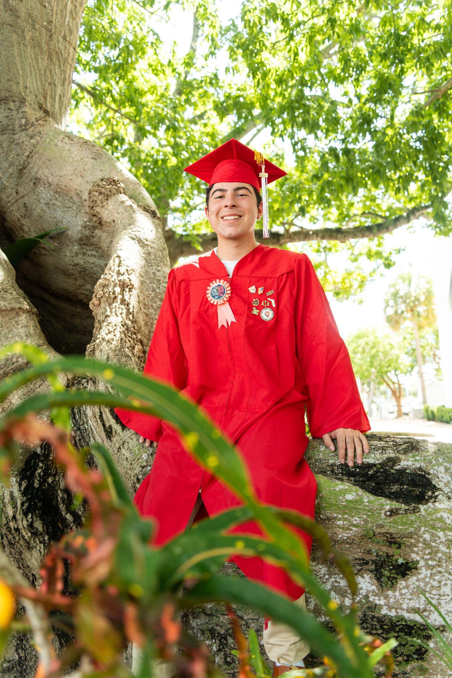 Smiling graduate in red cap and gown with honor medals poses by a large tree outdoors.
