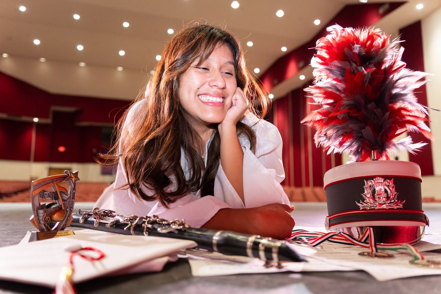 Smiling young woman in auditorium with marching band hat, clarinet, and sheet music on table.
