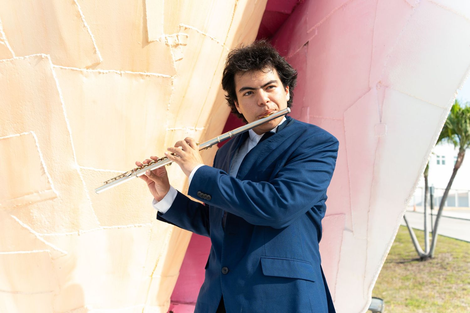 Young man in blue suit playing flute outdoors against colorful modern building wall.