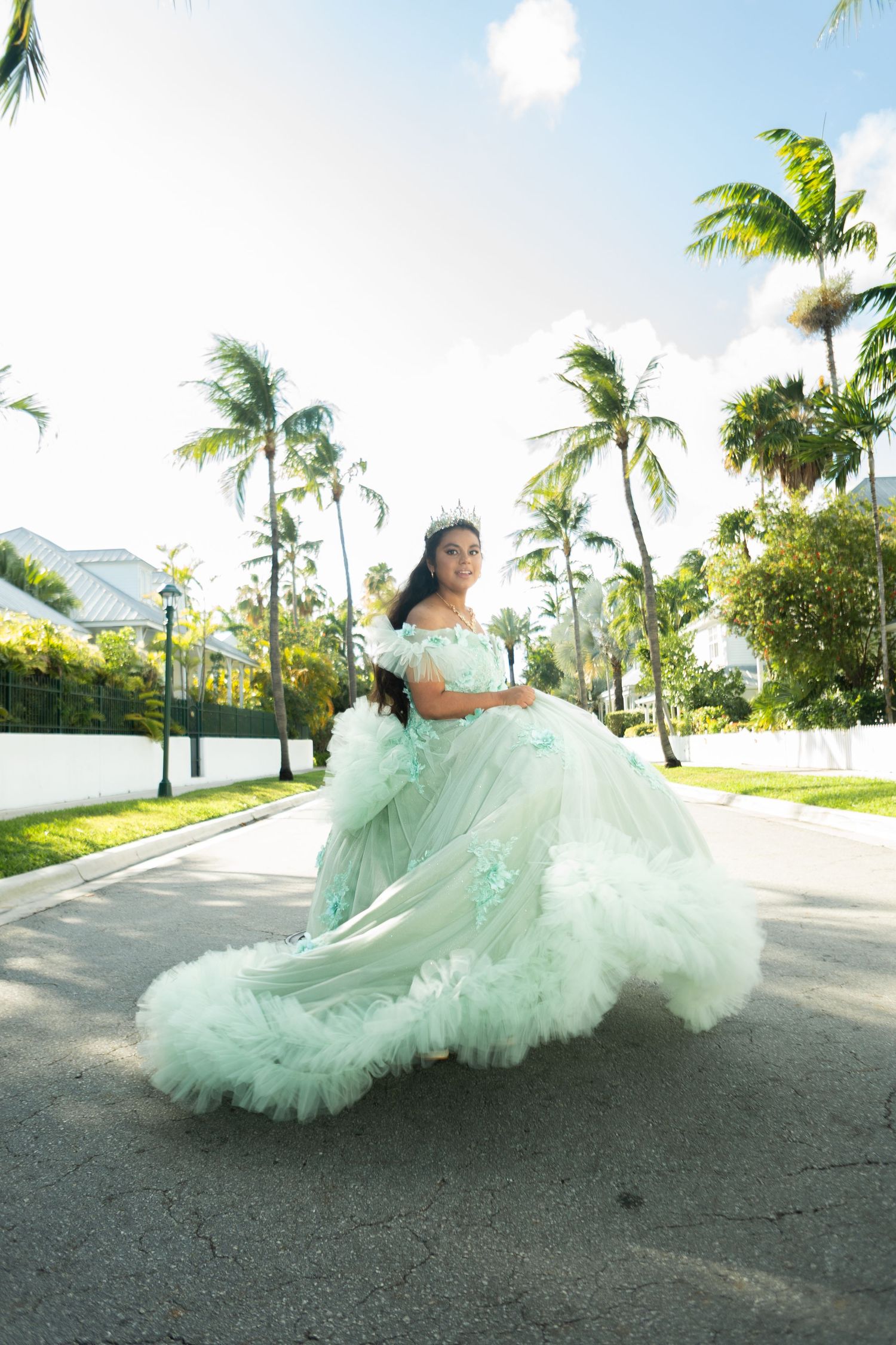 Young woman in mint green quinceañera ball gown twirling on a palm-lined street in a tropical setting.