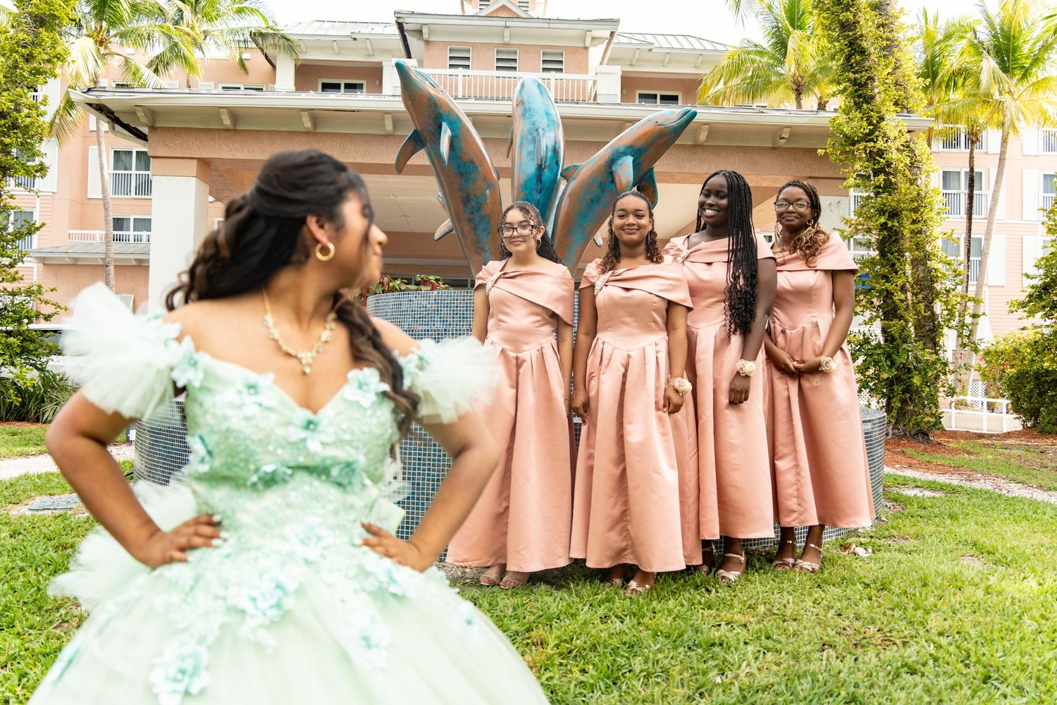 Quinceañera girl in mint green gown poses with bridesmaids in pink dresses outdoors near tropical garden.