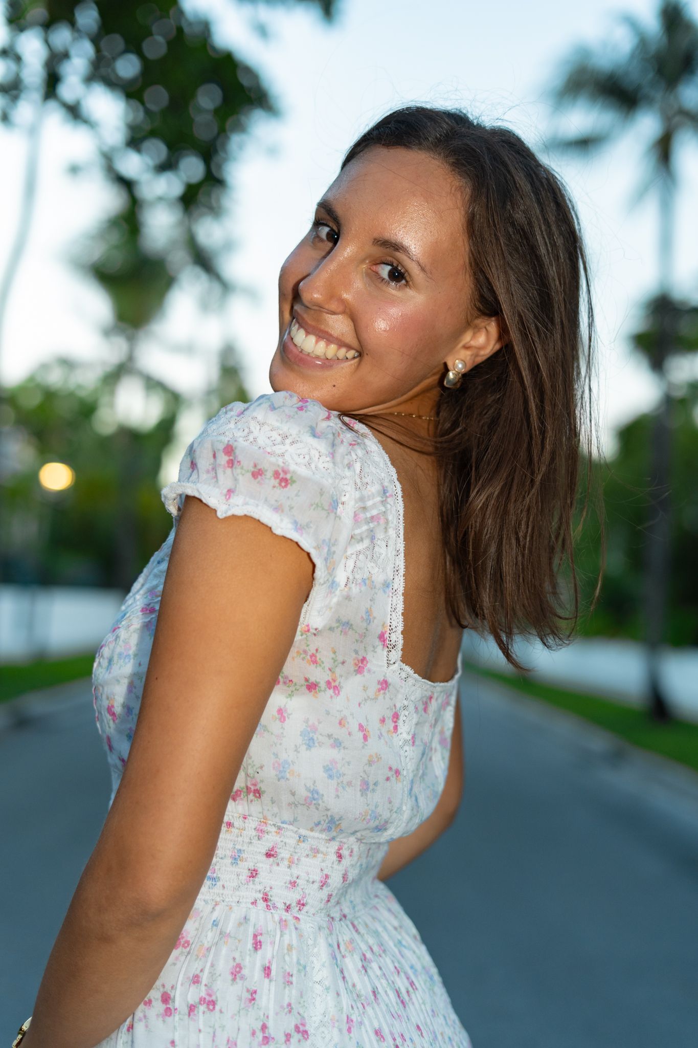 A person in a white floral dress poses playfully on a palm tree-lined street at dusk.