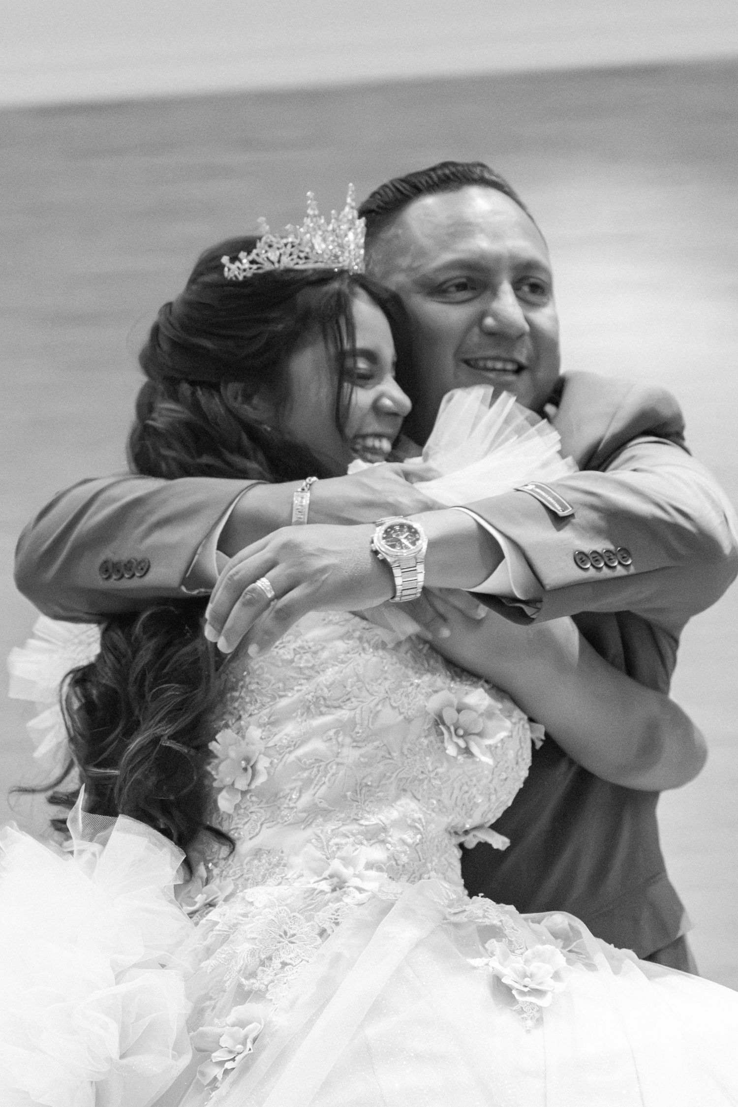 Black and white photo of a smiling bride in a tiara and groom sharing a joyful embrace at their wedding.