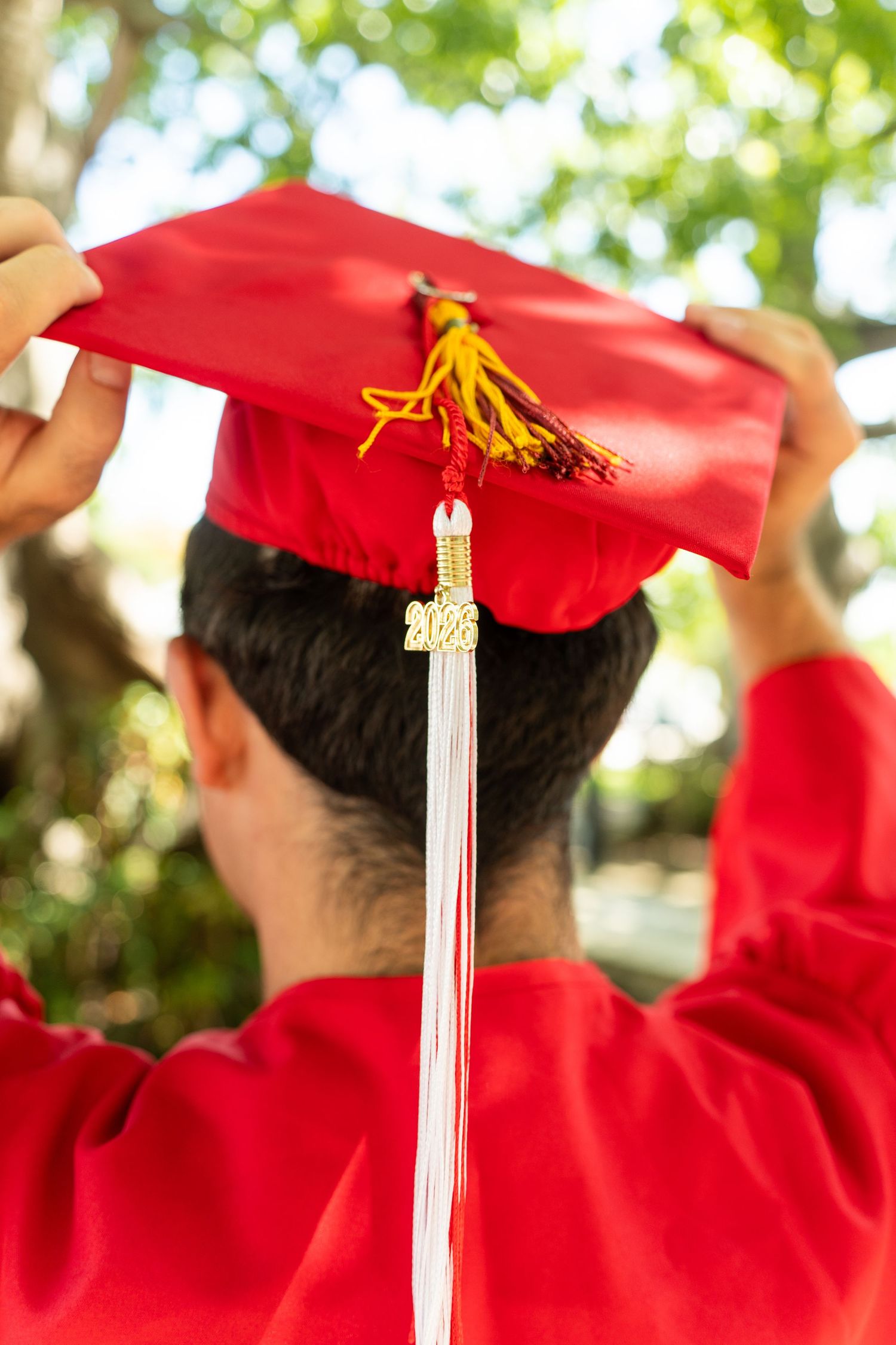 Graduate in red cap and gown adjusts mortarboard with gold tassel outdoors during graduation ceremony.