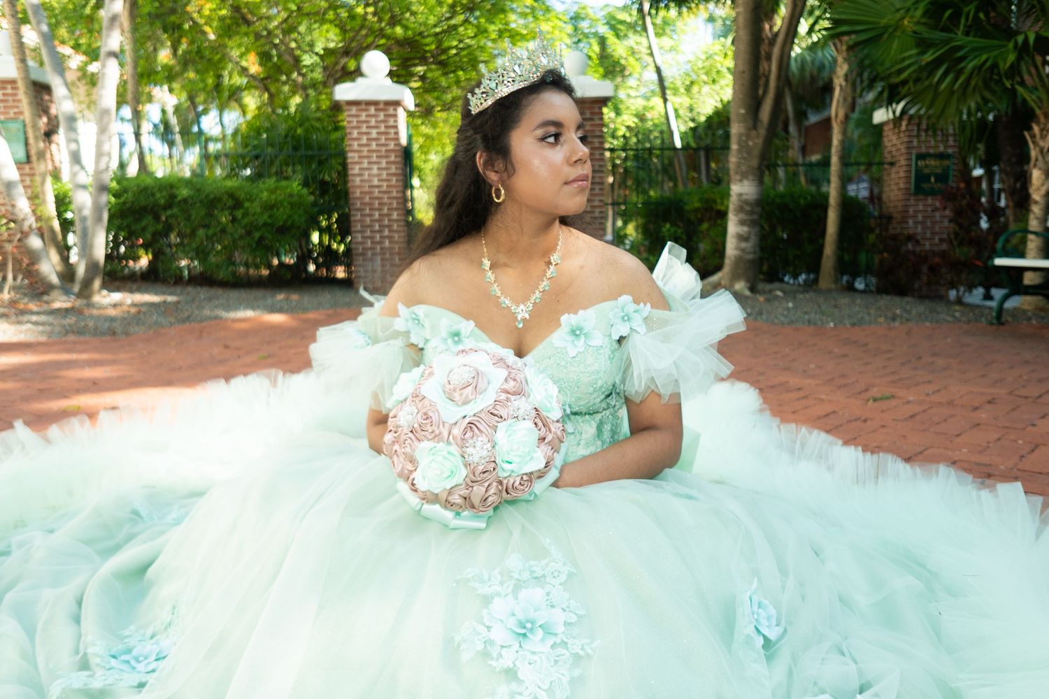 Young Latina girl in mint green quinceañera ball gown with tiara holds floral bouquet outdoors.