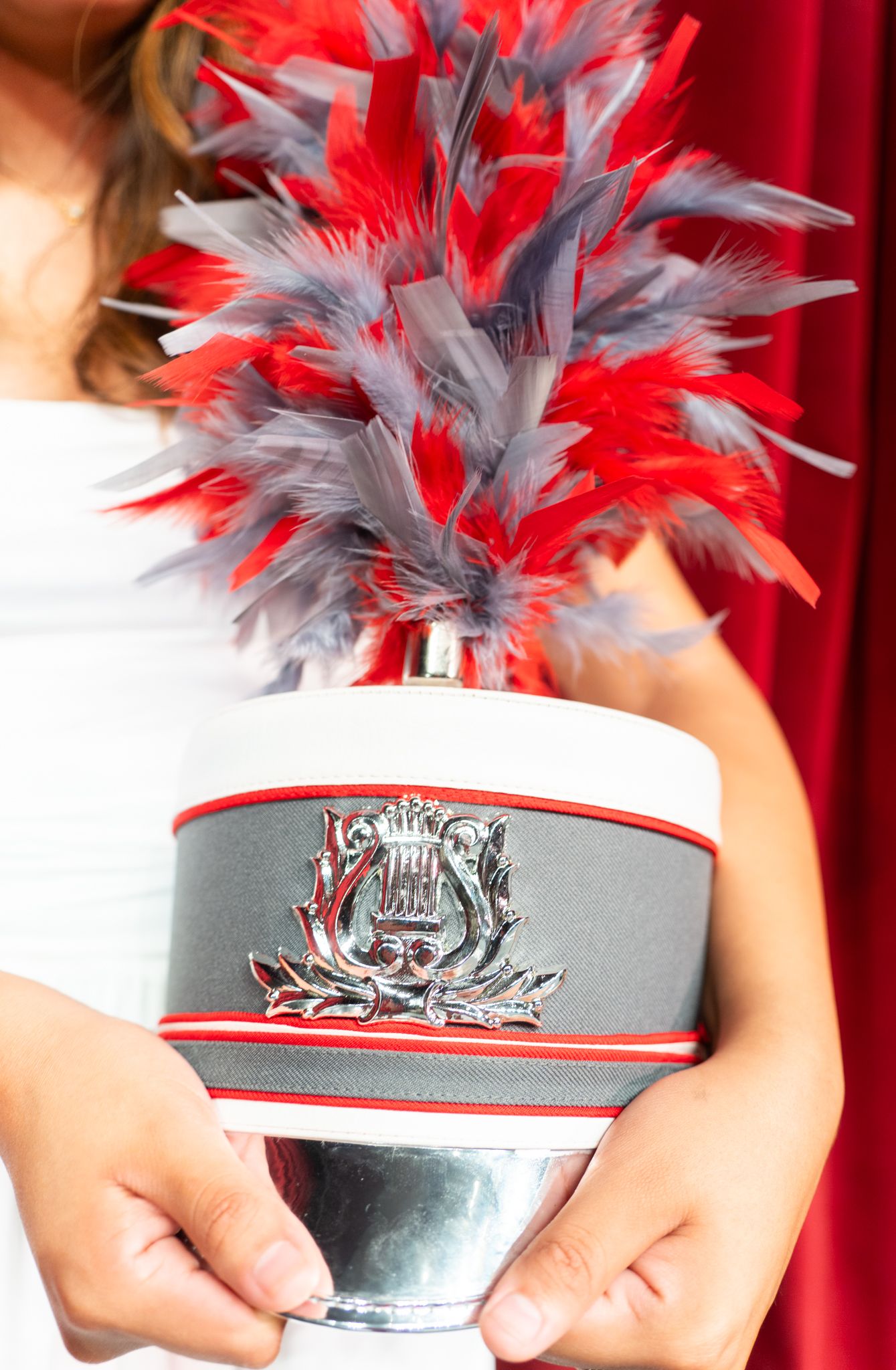 White marching band shako hat with silver badge and red and gray feather plume held by a person in white.