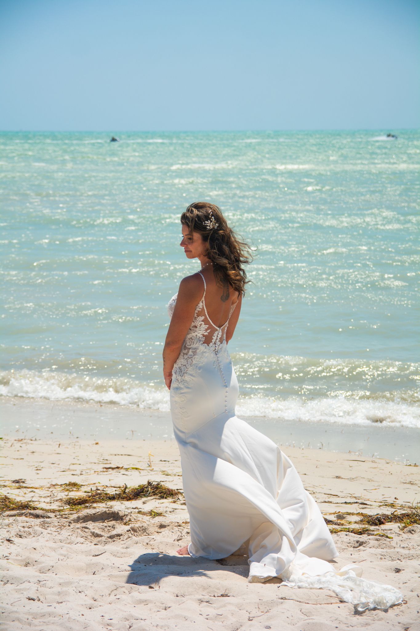 A bride in a flowing white dress stands on a beach looking out at turquoise waters.