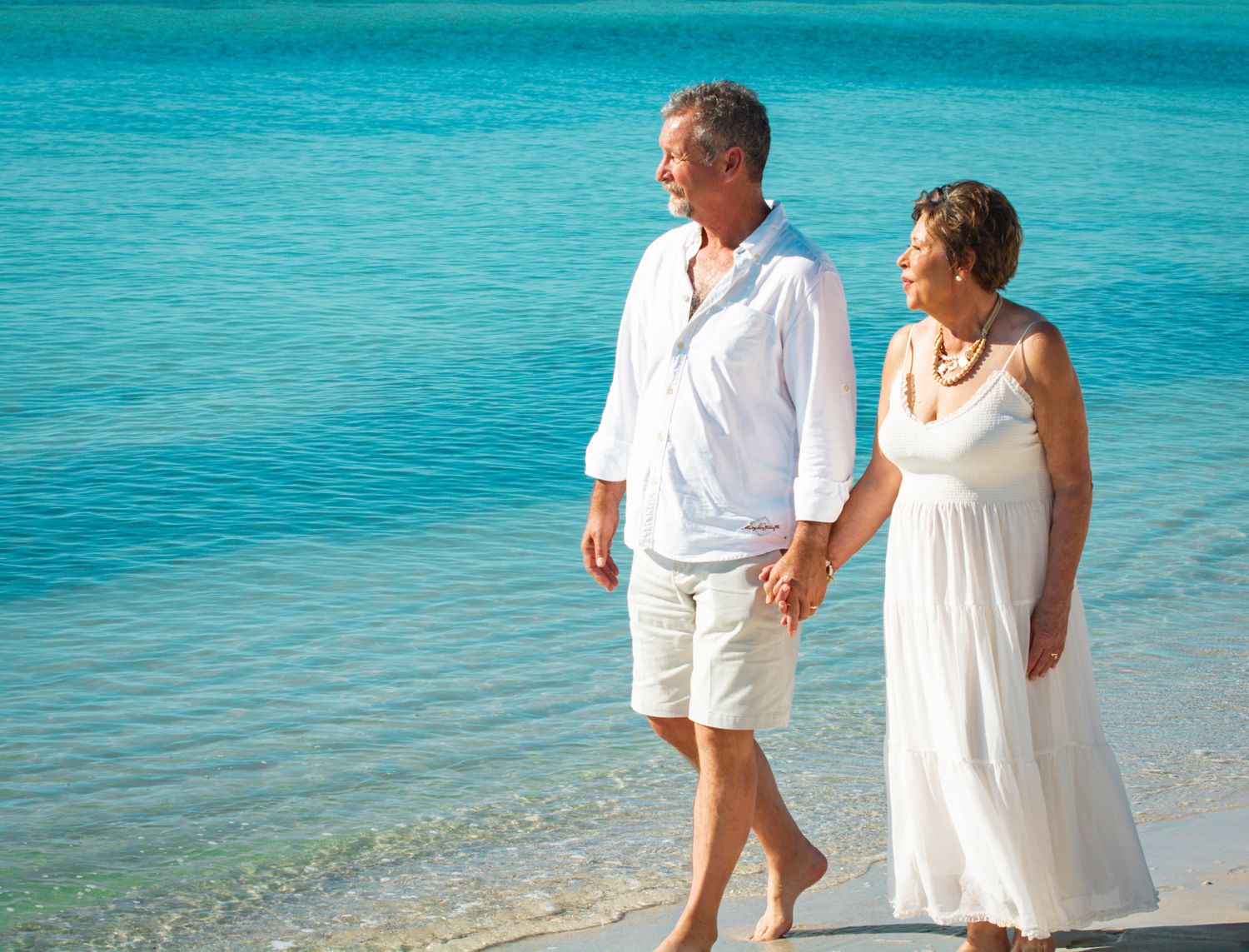 Couple walks hand in hand on tropical beach with turquoise water.