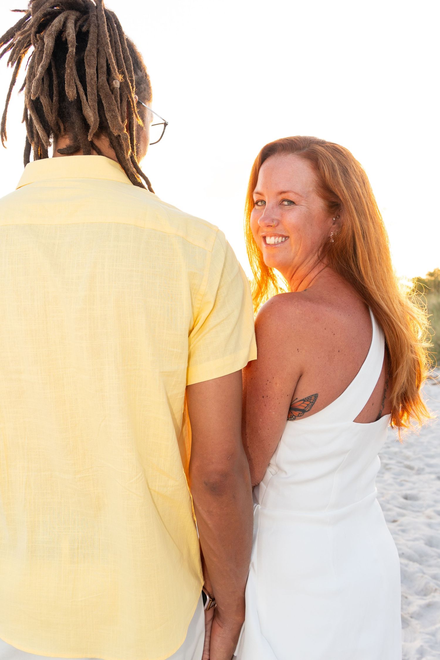 Smiling redhead woman in white dress holding hands with man in yellow shirt during beach photo session.
