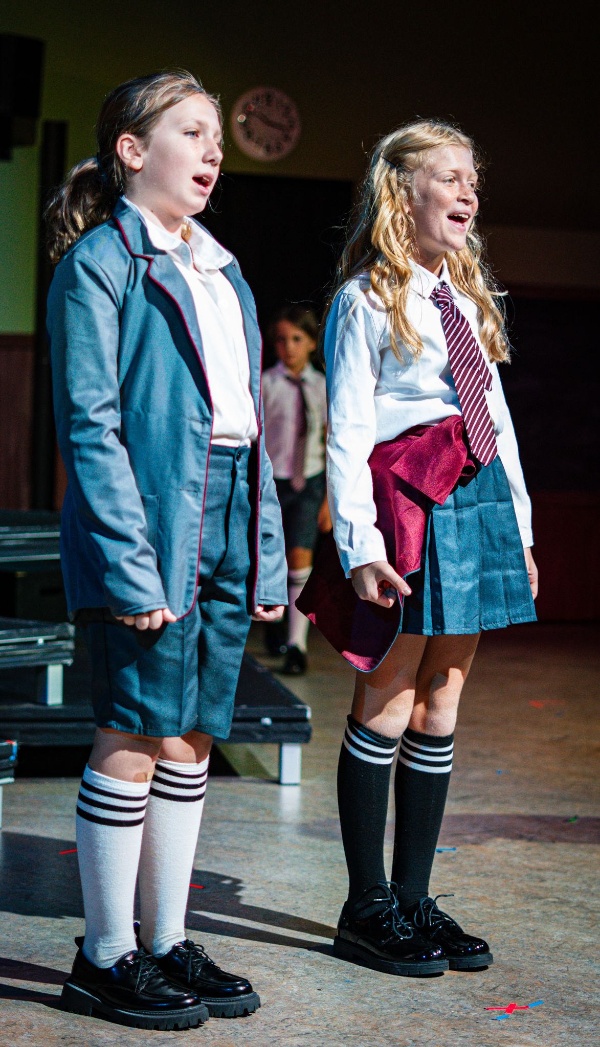 Two performers in school uniforms and striped knee socks singing together on stage.