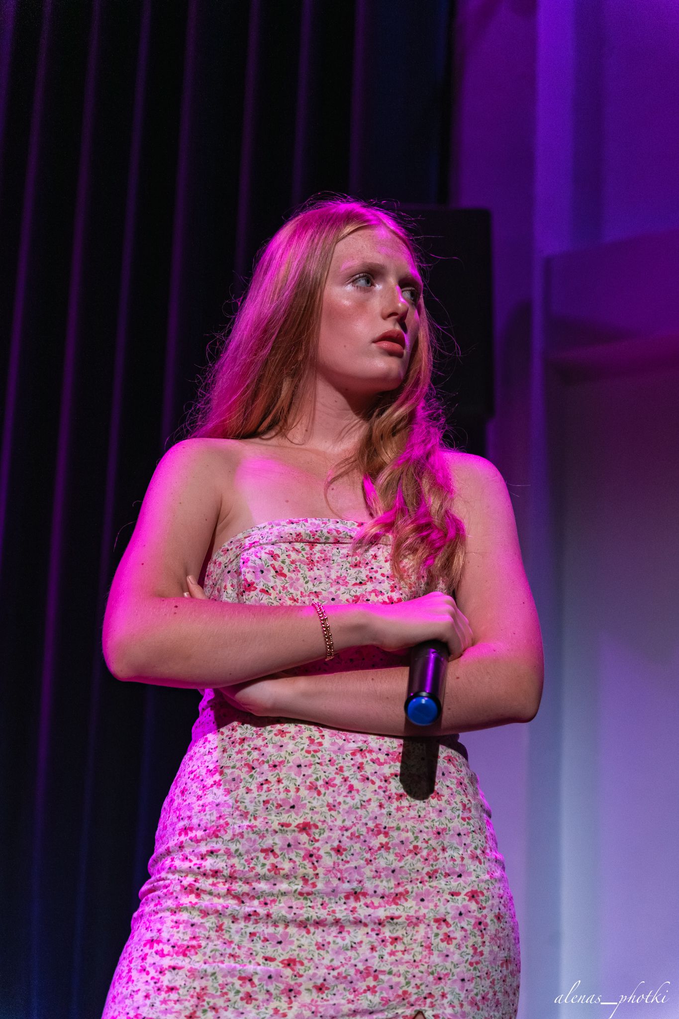 A performer in a floral dress sings on stage under dramatic purple and pink lighting during a concert performance.