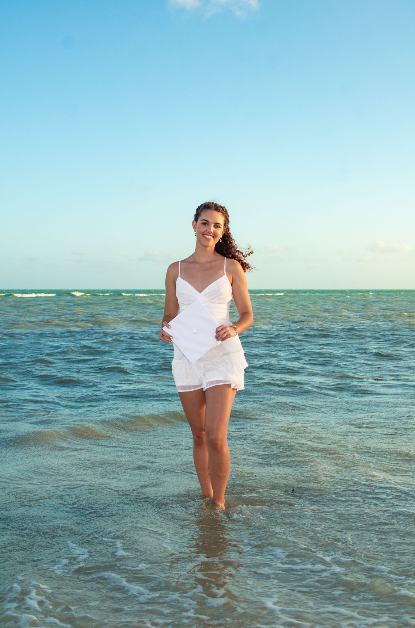 Person in white dress walking through turquoise ocean waters at beach.