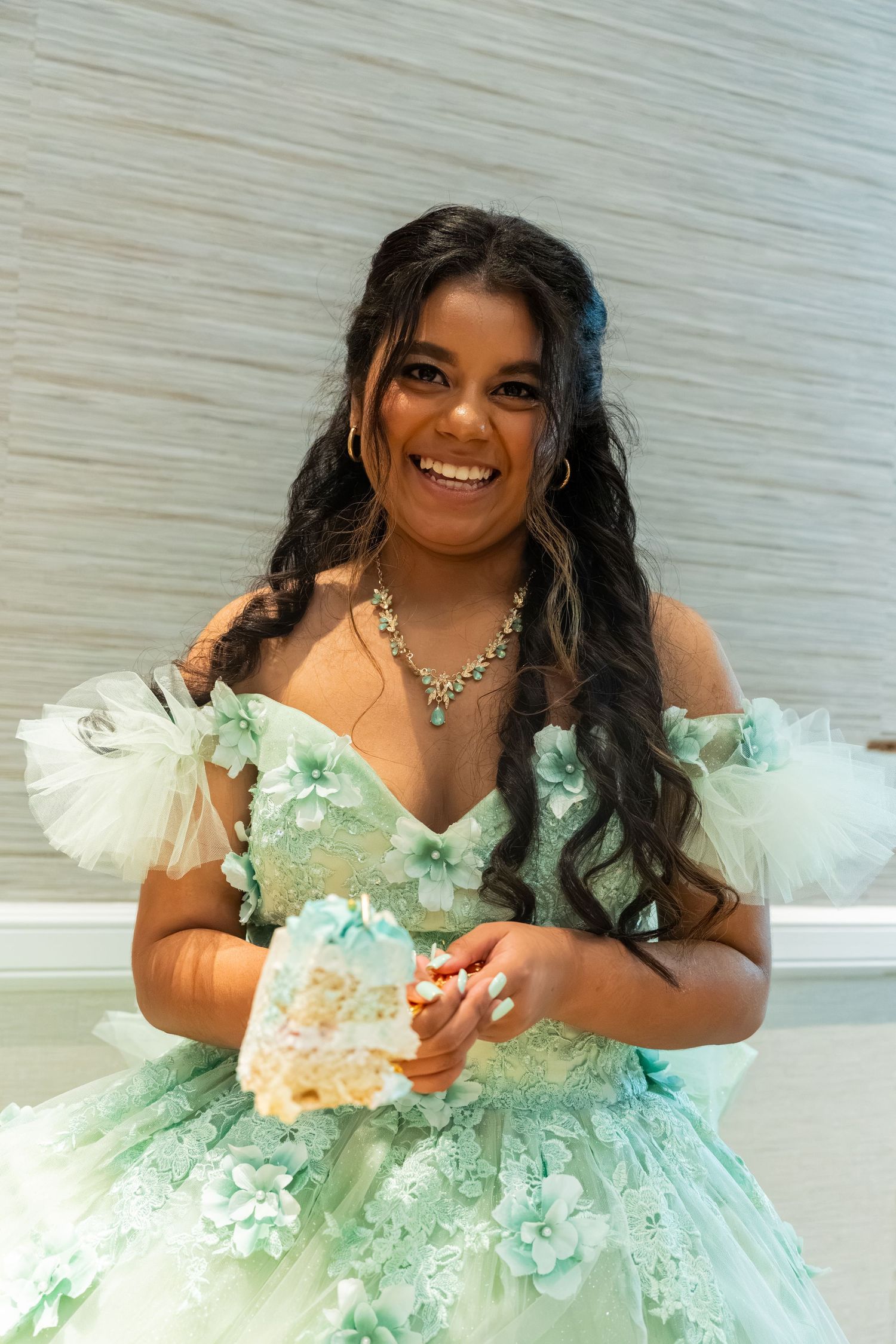 Smiling young woman in mint green floral quinceañera gown holding a slice of cake at her party.