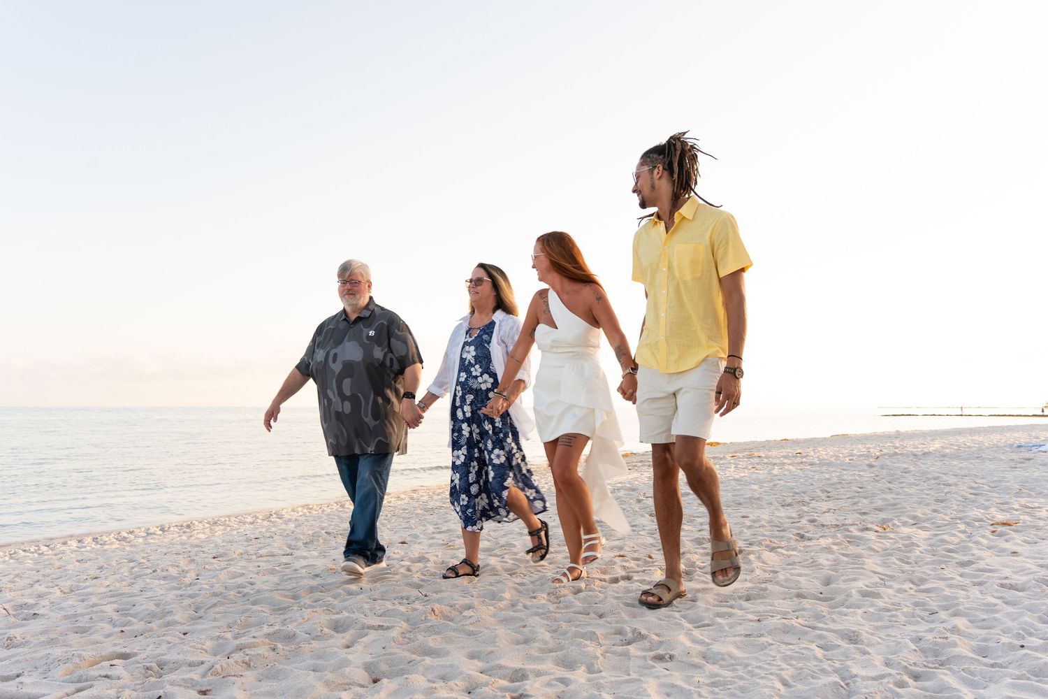 Two couples walk hand-in-hand along a sunny beach at sunset, enjoying a relaxing seaside stroll together.