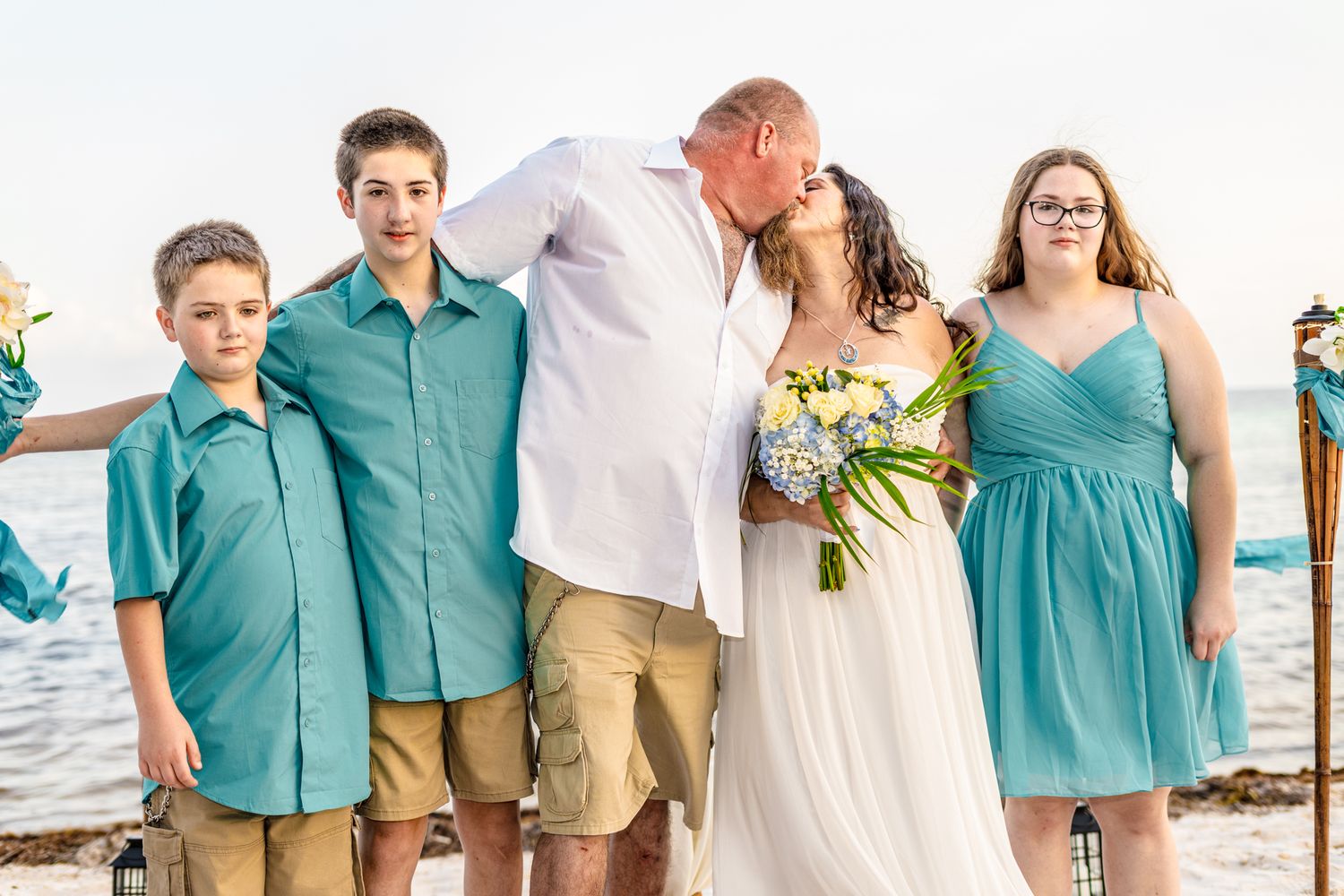 A beach wedding family photo with attendees wearing coordinated turquoise and white outfits.