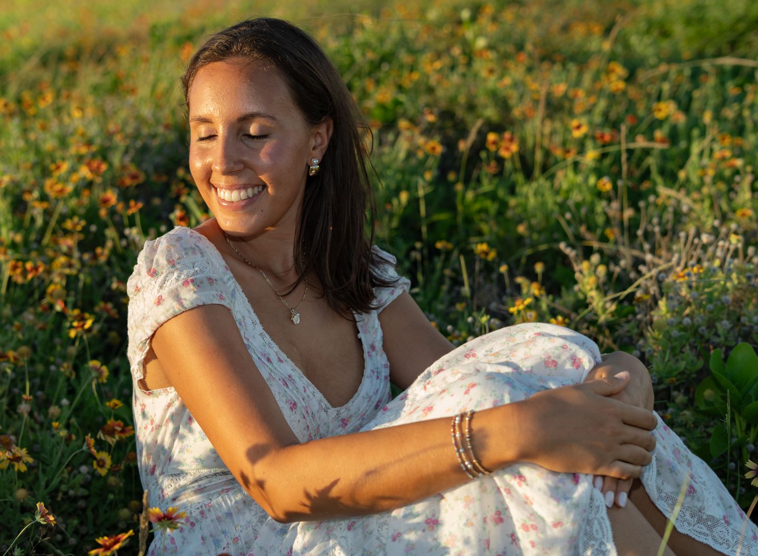 Someone in a white lace dress sits in a golden wildflower field at sunset with warm sunlight illuminating the scene.