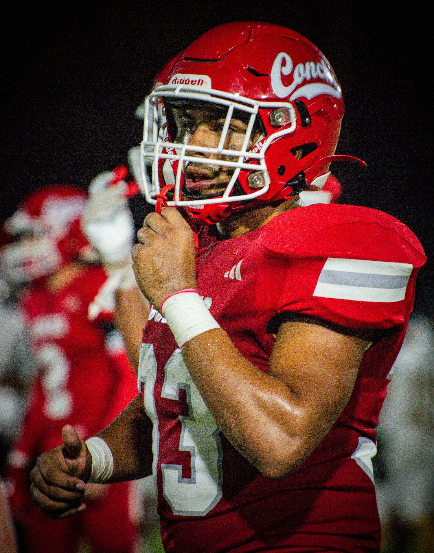 Football players in red helmets and uniforms prepare on the sideline under stadium lights.