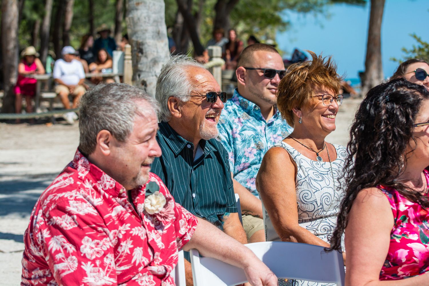 Wedding guests in colorful Hawaiian shirts and summer attire seated outdoors under palm trees.
