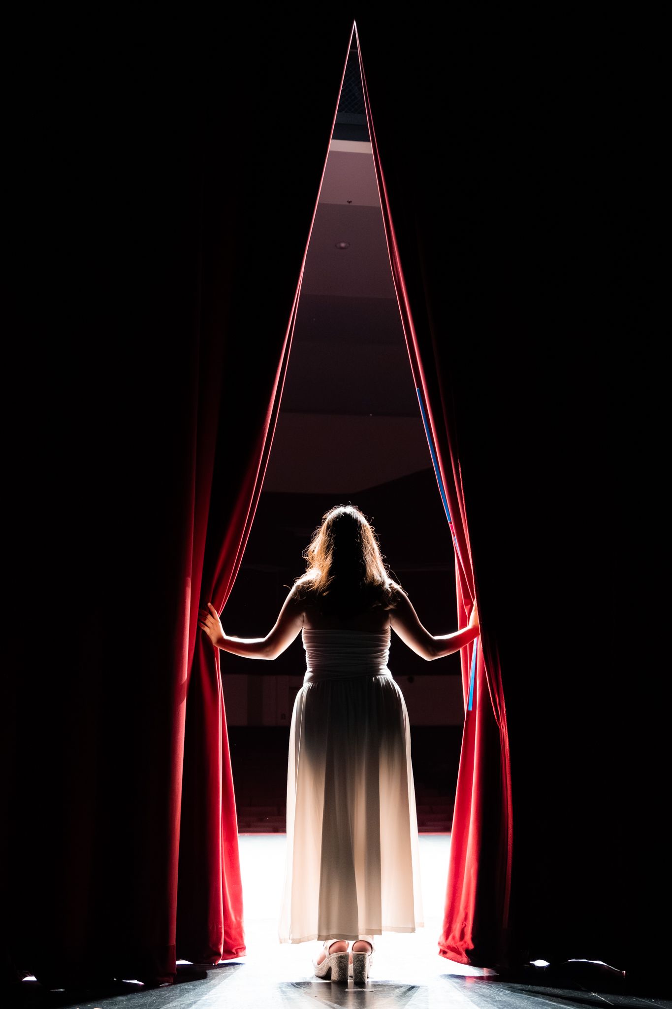 Woman in white gown dramatically parting red stage curtains with bright light streaming through the opening.