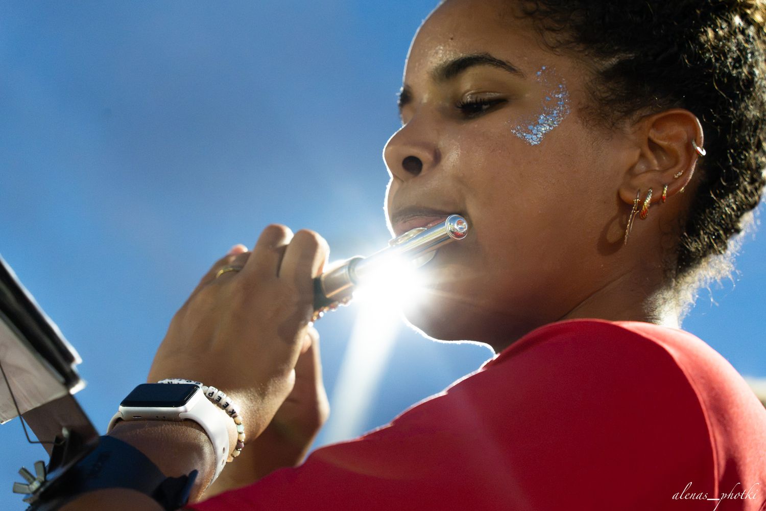 Woman in red top playing a silver flute outdoors under bright sunlight with blue sky background.