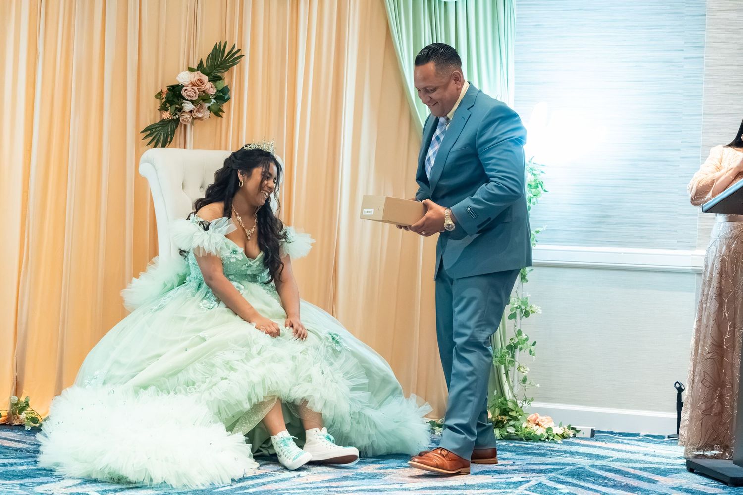 A young woman in a mint green quinceañera gown receives her ceremonial shoes from a man in a blue suit.
