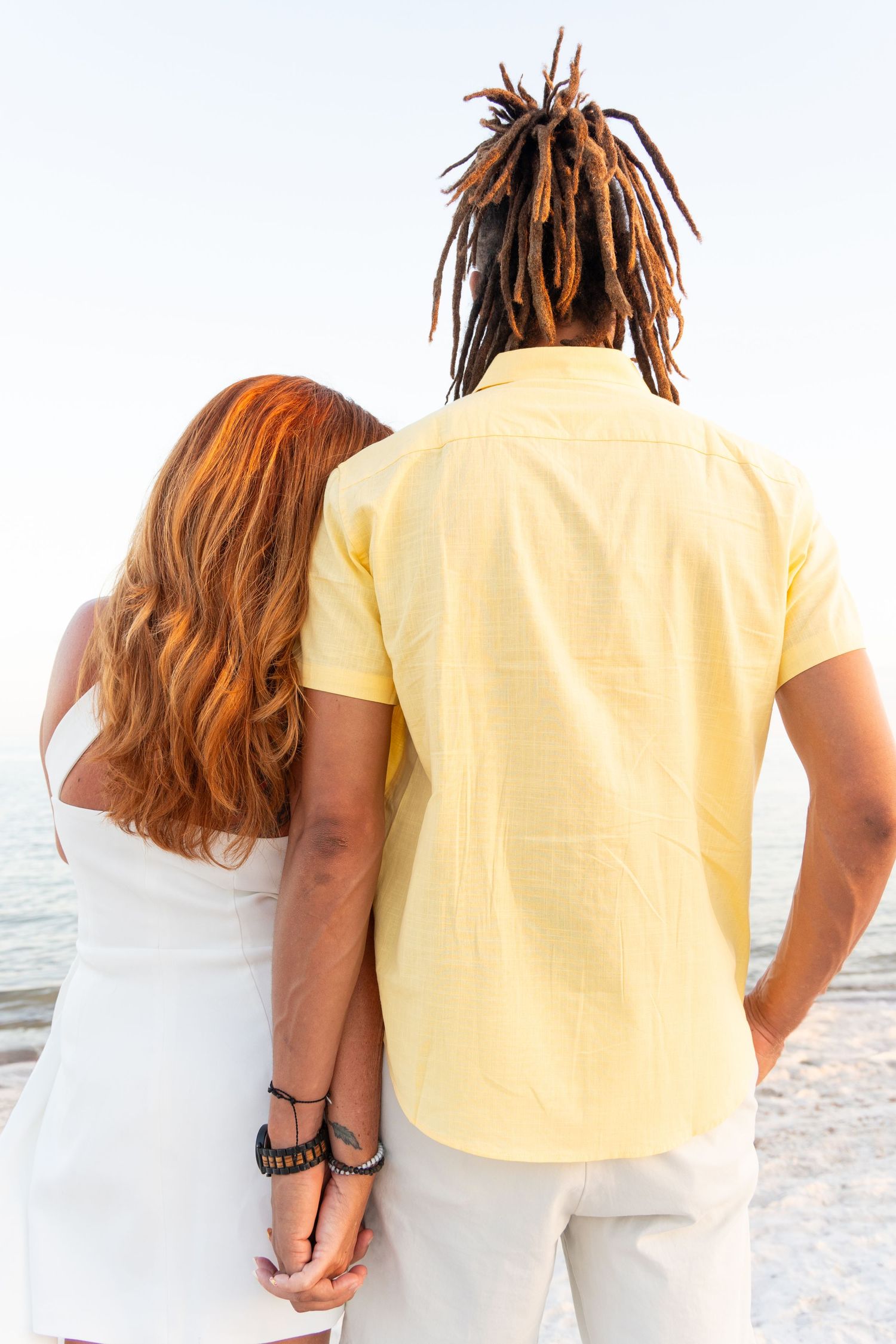 Couple standing arm in arm from behind, looking at the ocean during a romantic beach engagement session.