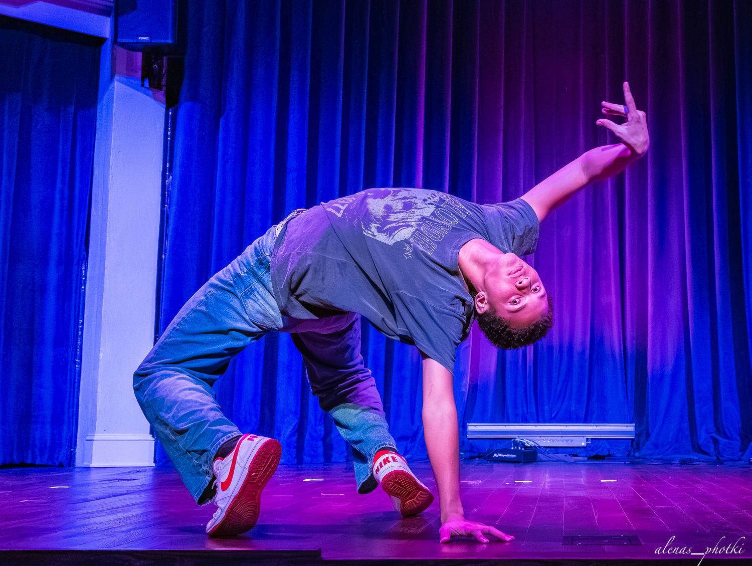 Dancer performs dynamic breakdance moves on stage illuminated by dramatic blue and purple lighting.
