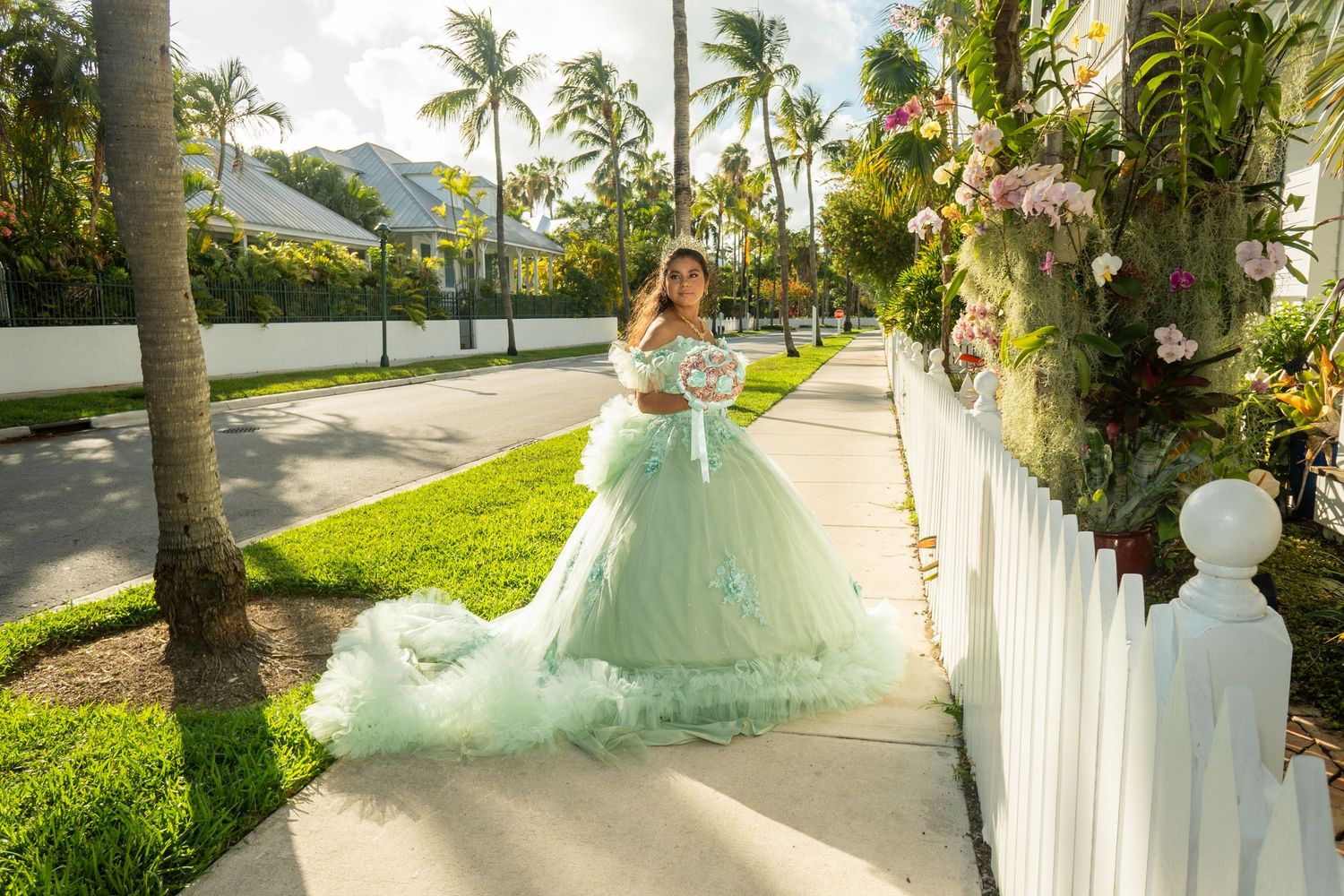 Young woman in a mint green ball gown quinceañera dress poses on a sunny sidewalk lined with palm trees.