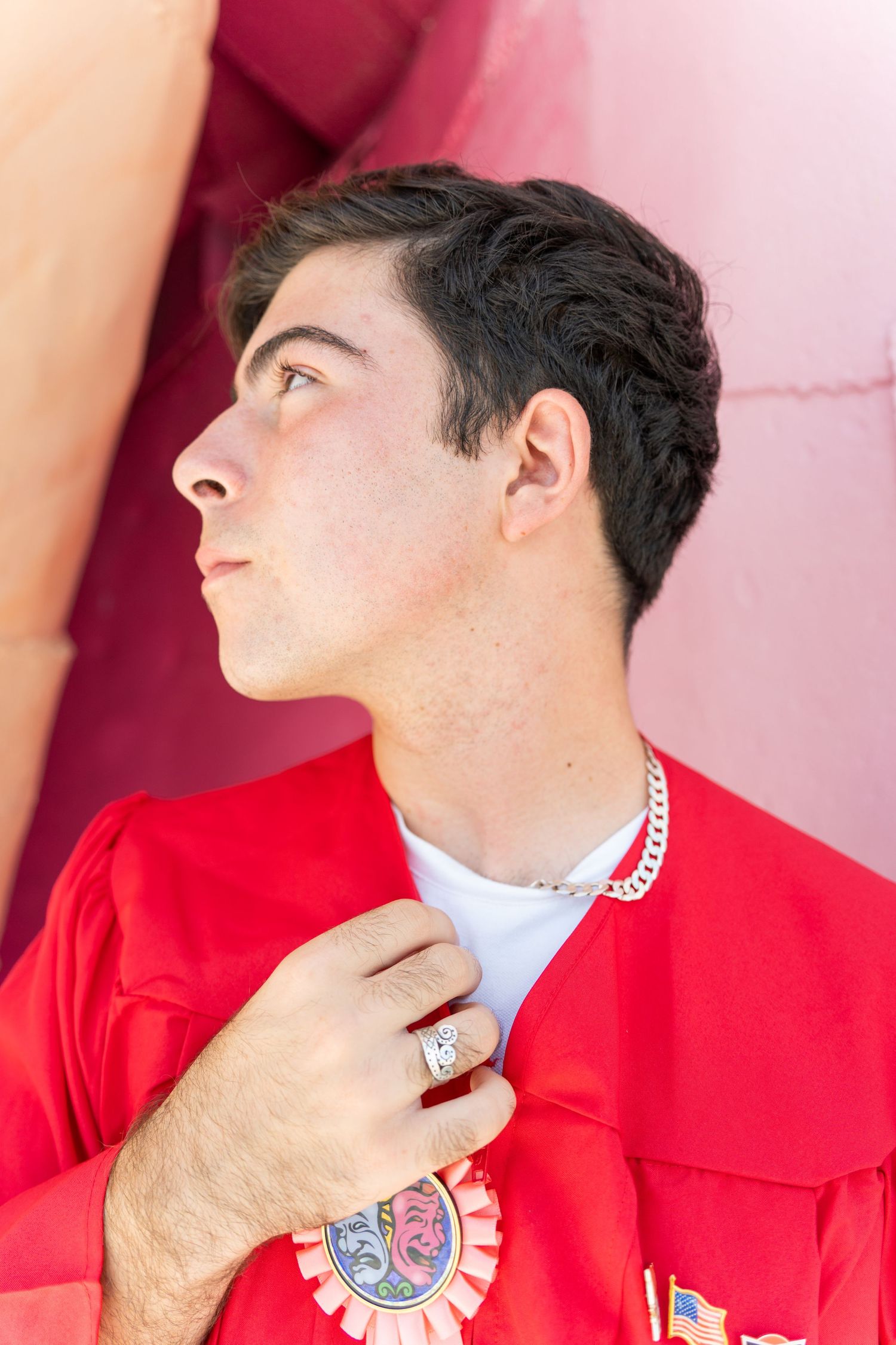 Young man in red graduation gown holding lapel, wearing silver chain necklace and ring, looking upward confidently.