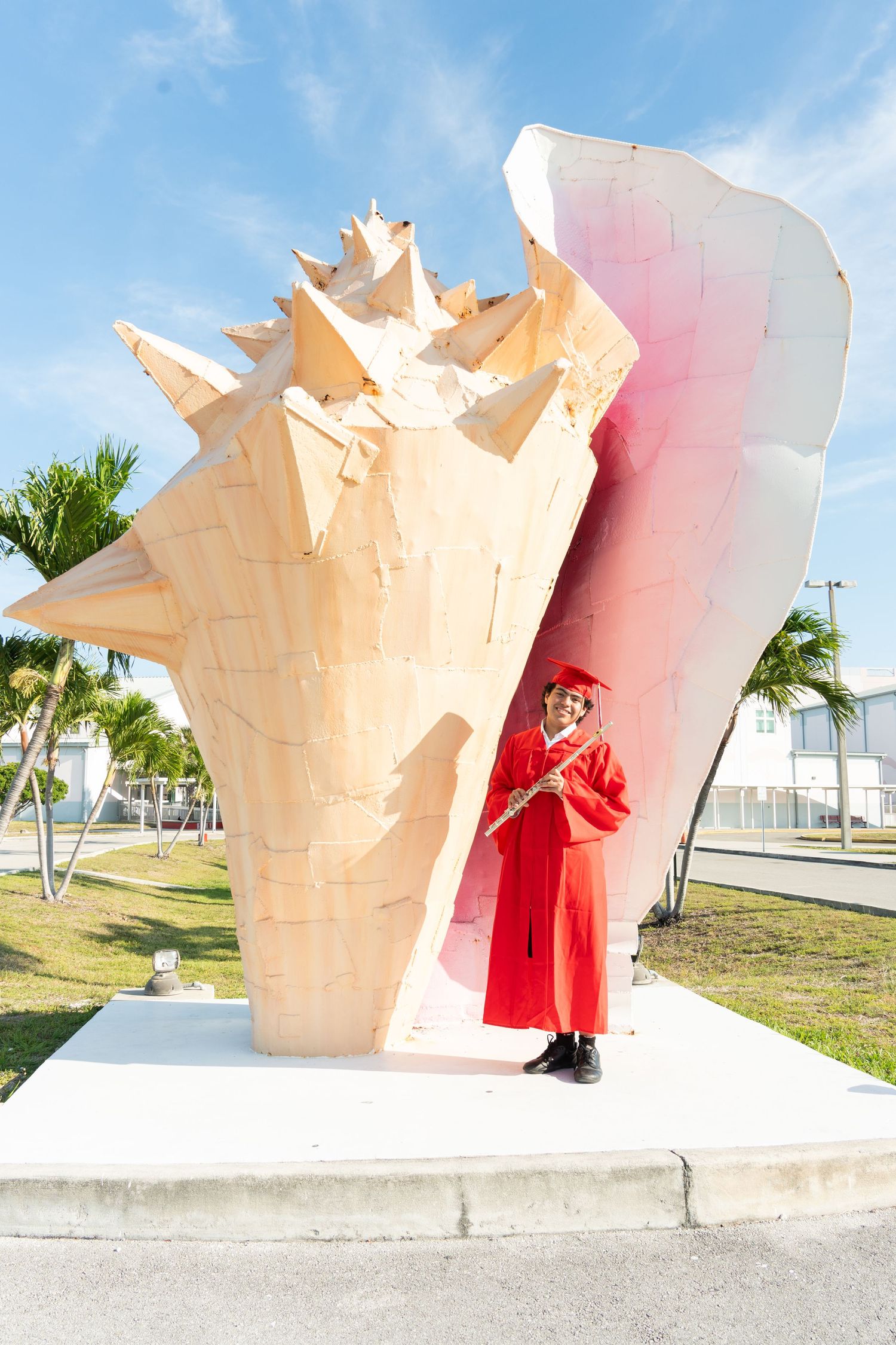 Graduate in red cap and gown poses with a large conch shell sculpture outdoors on a sunny day.