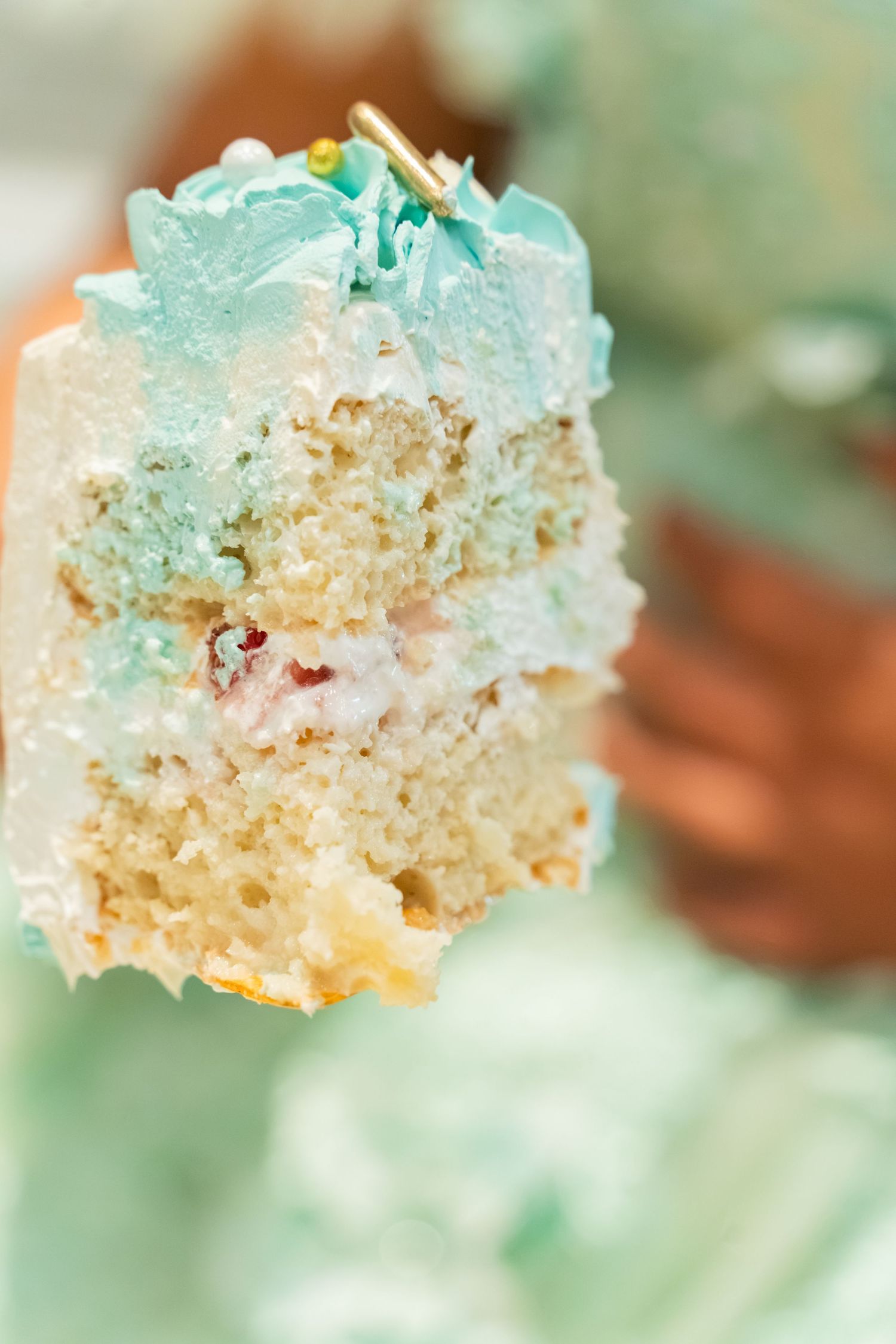 A slice of light blue frosted cake with white cream filling held up close, showing moist layers and decorative teal icing.