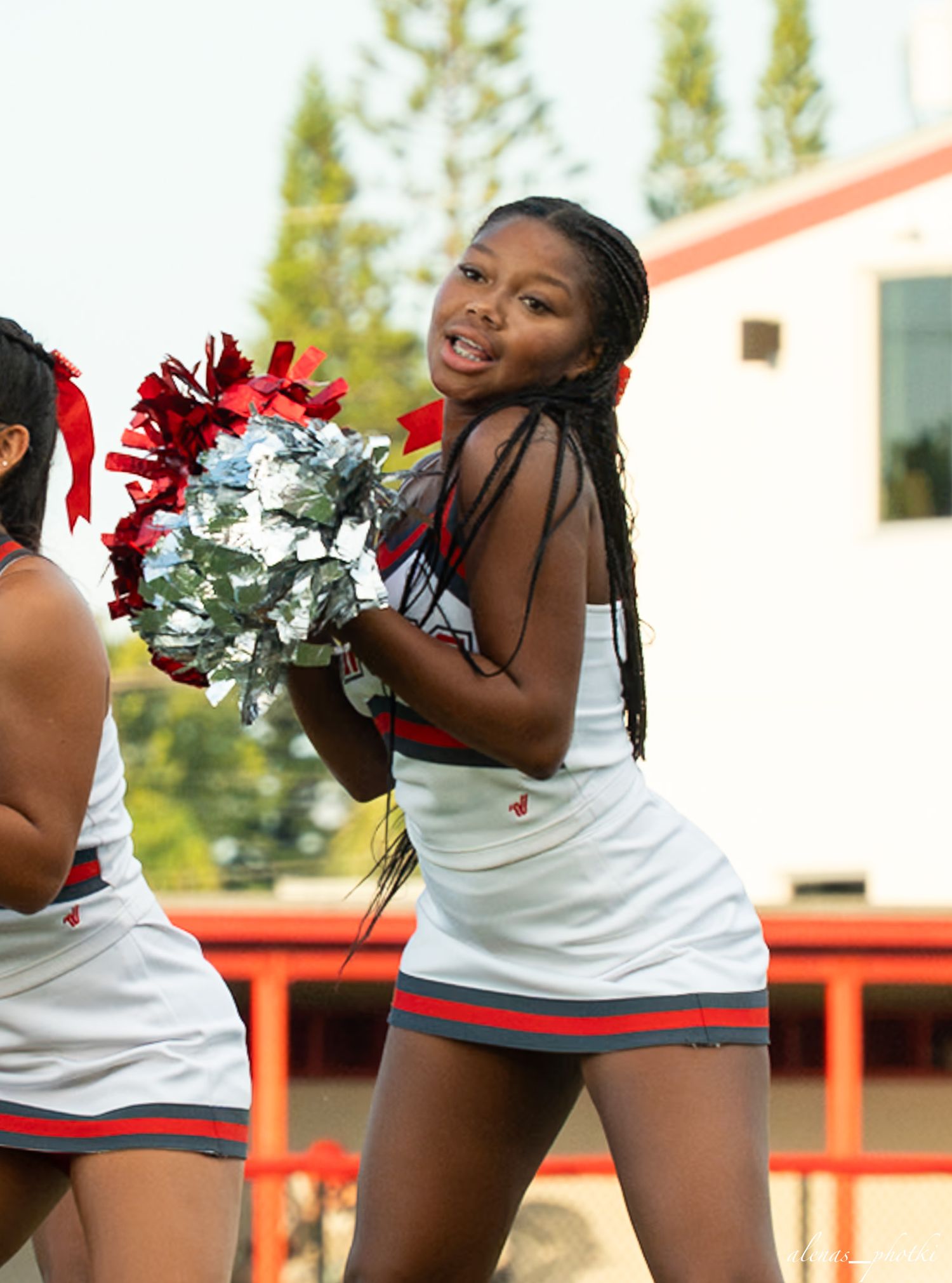 Young cheerleader in white and red uniform smiles while holding red and silver pom-poms during outdoor performance.
