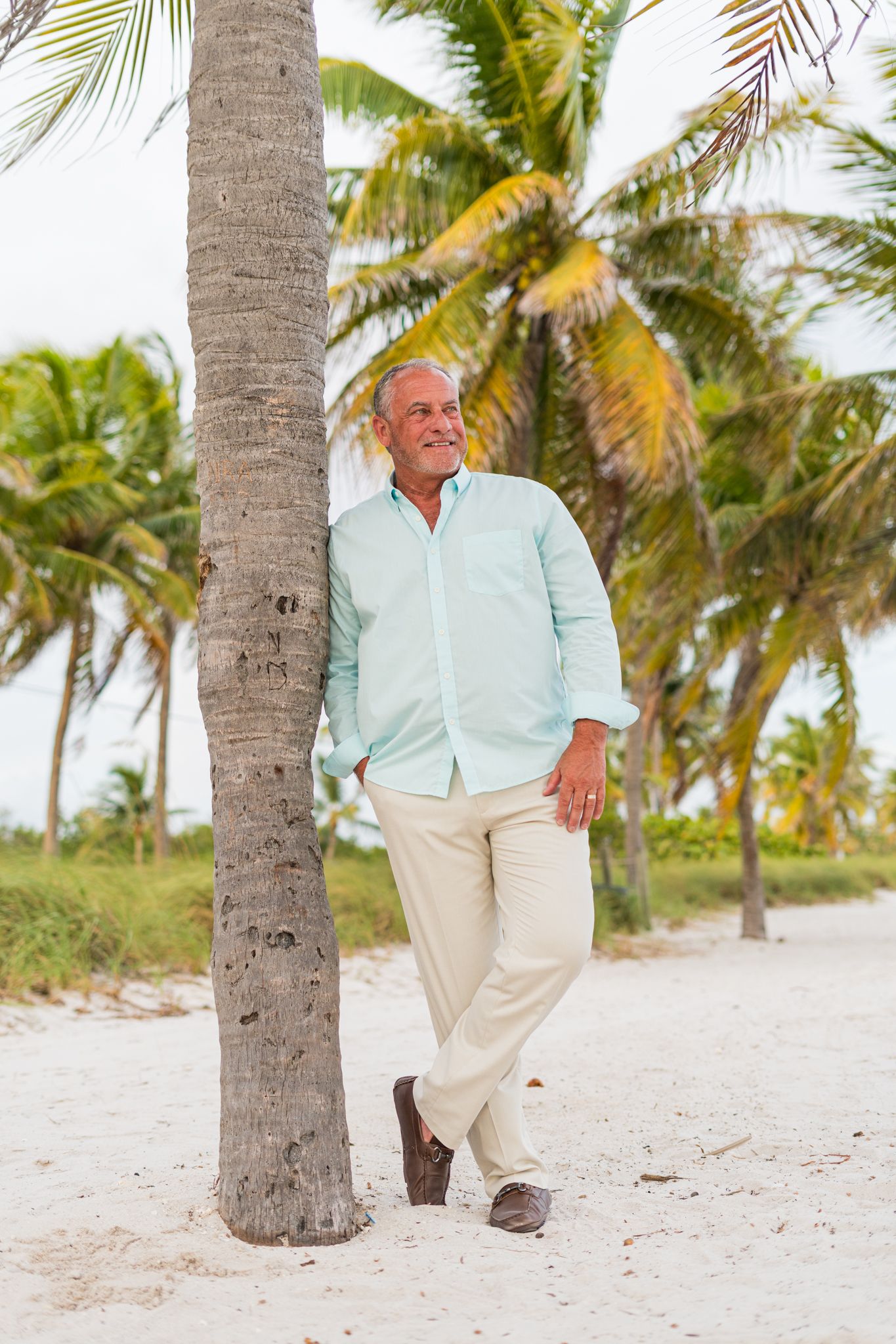 A person in casual beach attire leaning against a palm tree on a sandy tropical path.