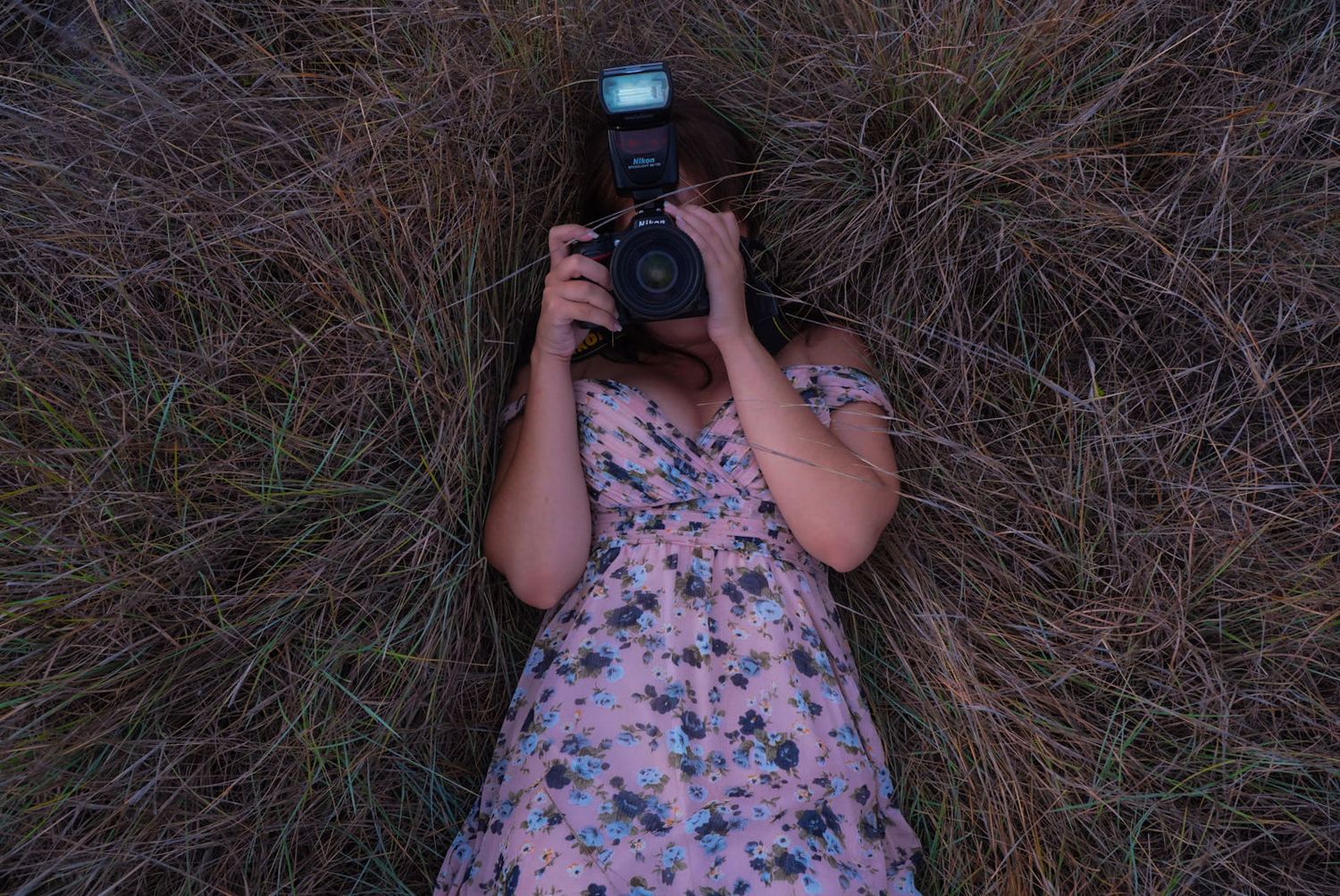 Figure in floral dress photographing in tall grass against blue sky.