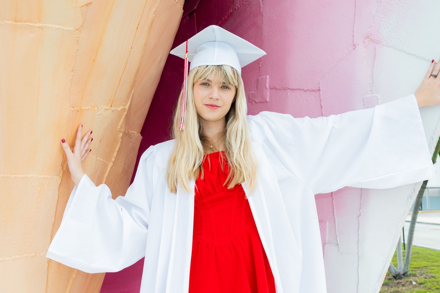 Young woman in white graduation cap and gown poses confidently outdoors against colorful background.