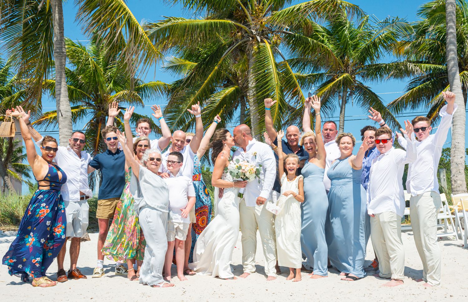 Wedding party celebrating with raised arms on a tropical beach with palm trees.