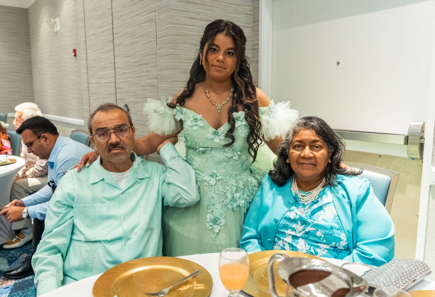 Young woman in mint green quinceañera gown poses with two seated adults at a celebration dinner table.
