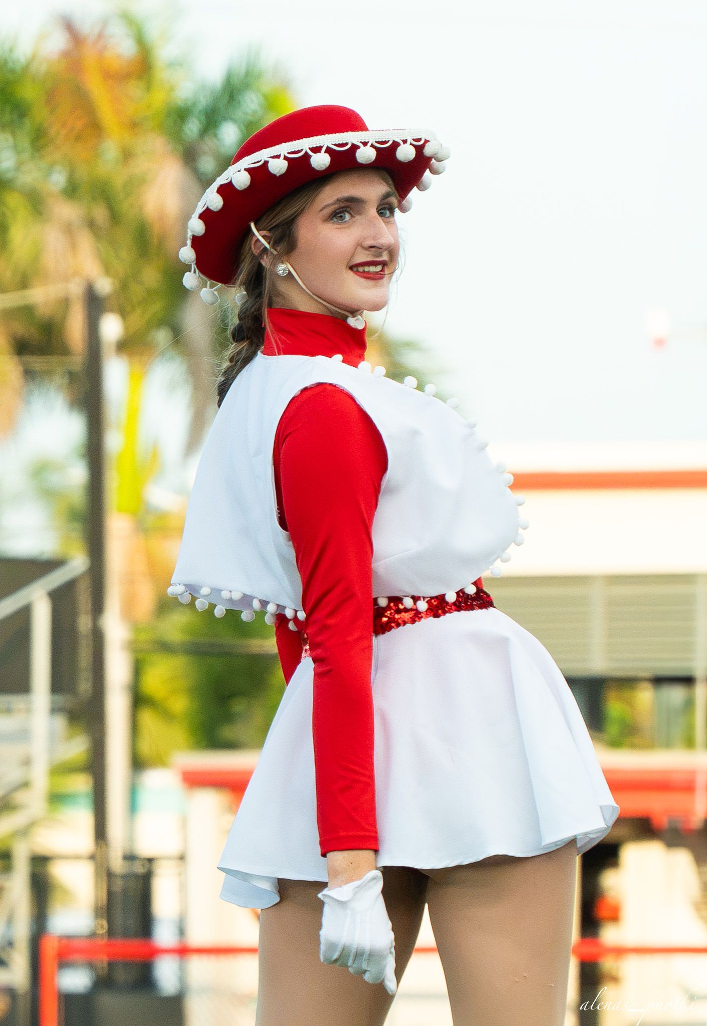 Young woman in red and white majorette costume with pom-pom hat smiling outdoors near palm trees.