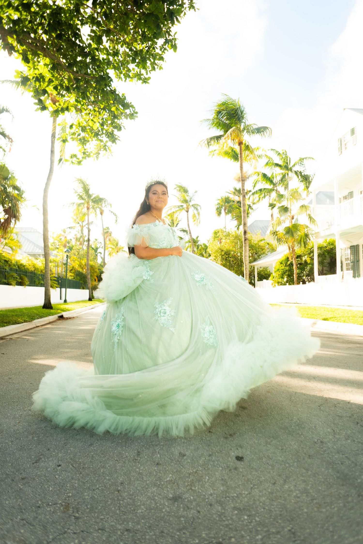 Young woman in a flowing mint green ball gown with floral details poses outdoors among palm trees.
