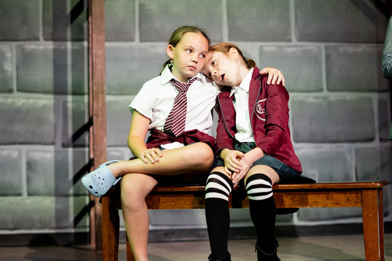 Two students in school uniforms sitting together on a wooden bench during a theatrical performance.