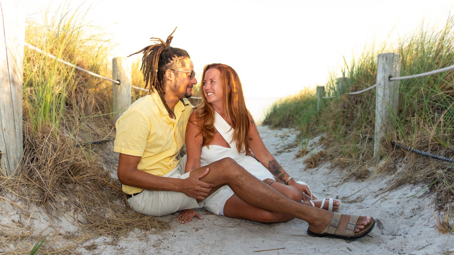A happy interracial couple sitting together on a sandy beach path, smiling and enjoying a sunny day outdoors.