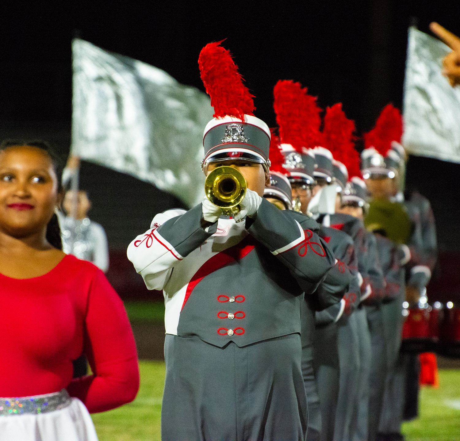 Marching band member plays trumpet in grey uniform with red plume during nighttime performance.