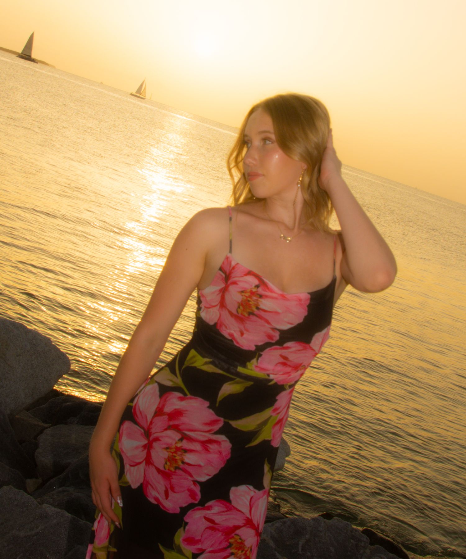 Woman in floral dress poses against sunset ocean view on rocky shore.