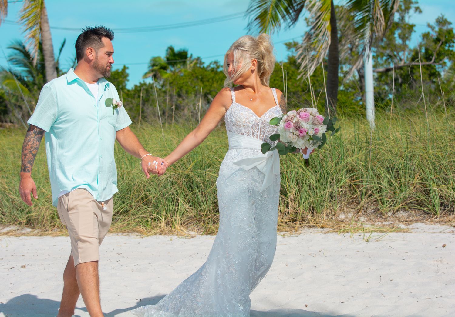 Couple walking on beach path holding hands with bride carrying purple bouquet.