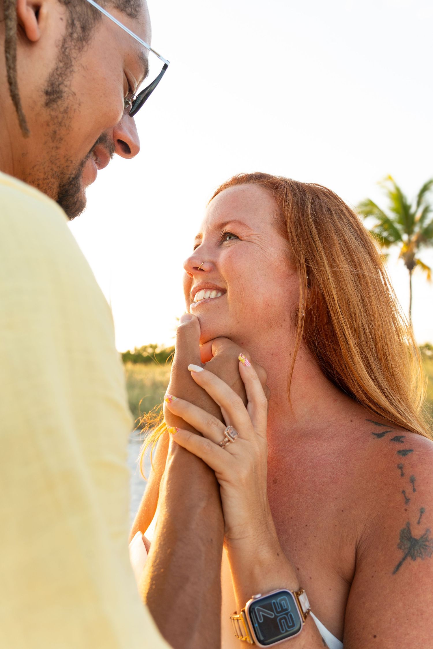 A smiling redhead woman holds hands with a man in a yellow shirt at a sunny beach, wearing an Apple Watch and ring.